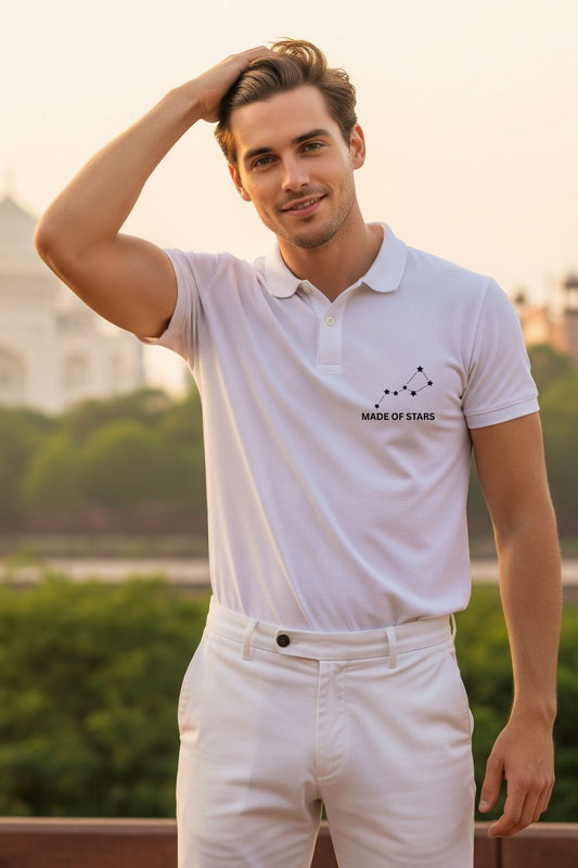 A stylish man wearing a ZUPPI 'Made of Stars' white polo t-shirt, showcasing the small constellation and text design on the front. He is seen with his fingers in his hair, in a relaxed, urban outdoor setting.