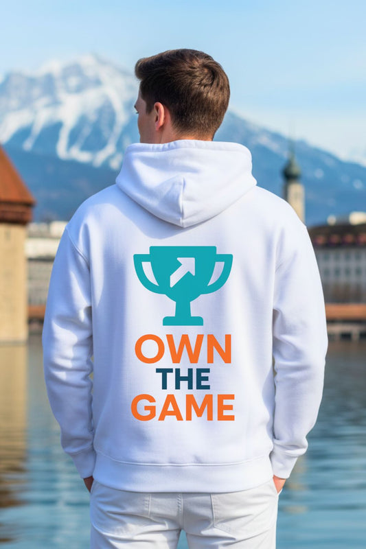 Back view of a man wearing a white oversized hoodie with 'Own the Game' text and logo, standing by a body of water with mountains in the background.
