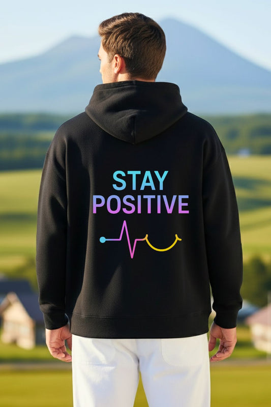back view of a man wearing a black oversized hoodie with 'Stay Positive' text in colorful letters, standing outdoors with a scenic background.