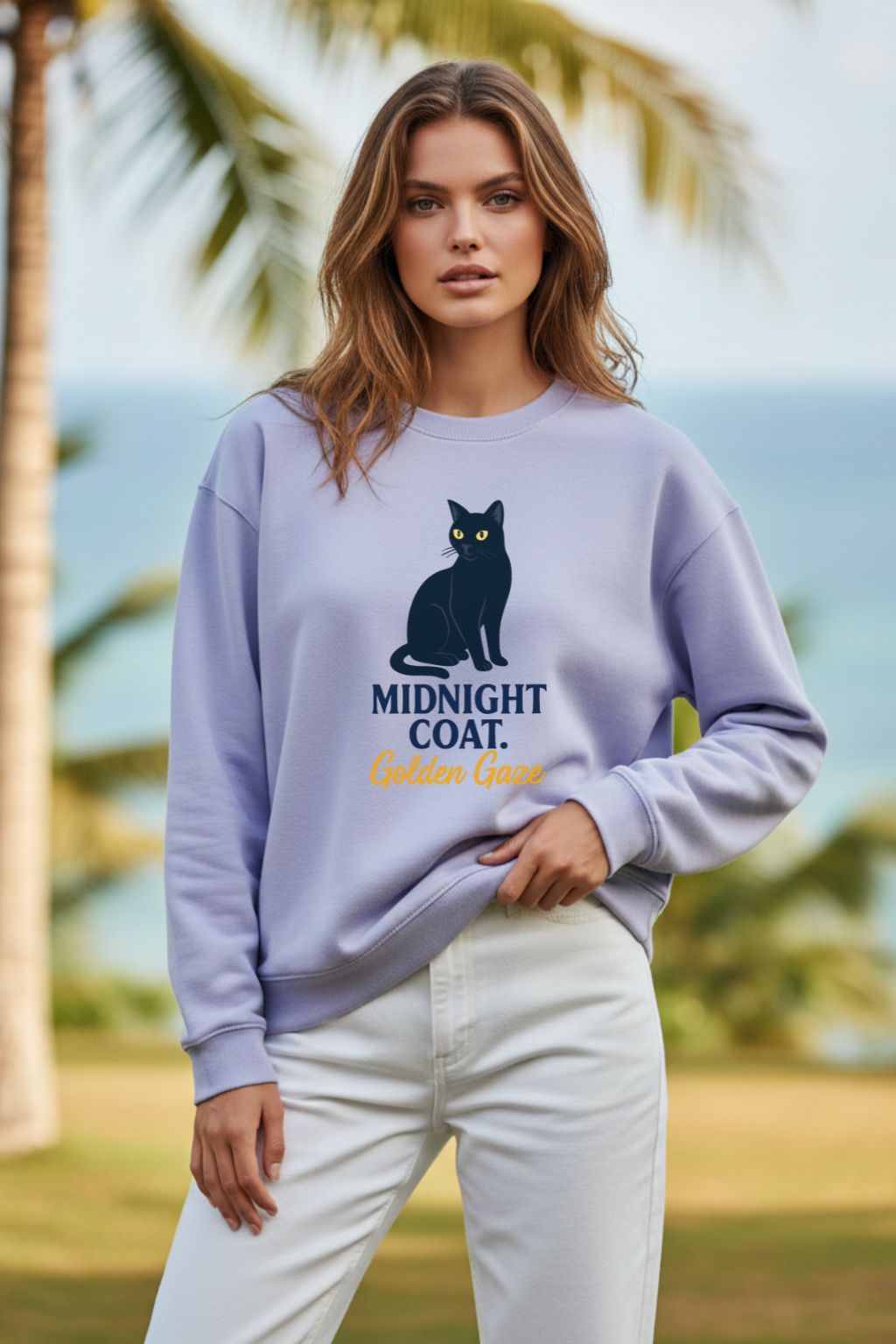 Woman wearing a light purple oversized sweatshirt with a cat graphic and text, standing outdoors with palm trees and ocean in the background.