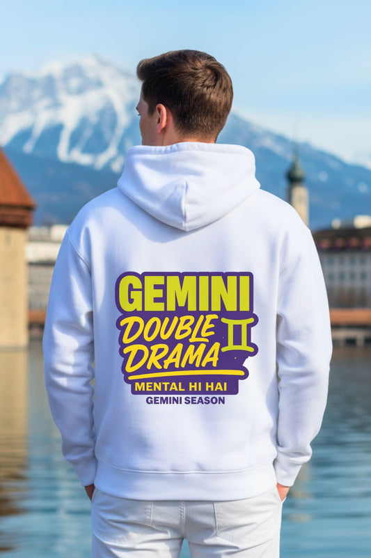 Man wearing a white hoodie with 'Gemini Double Drama' text, standing by a body of water with mountains in the background.