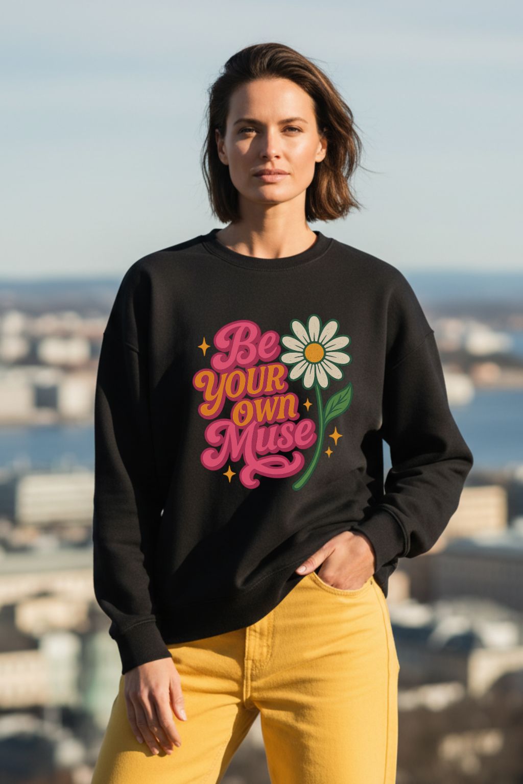 Woman wearing a black oversized sweatshirt with 'Be Your Own Muse' text and flower graphic, standing against an outdoor background.