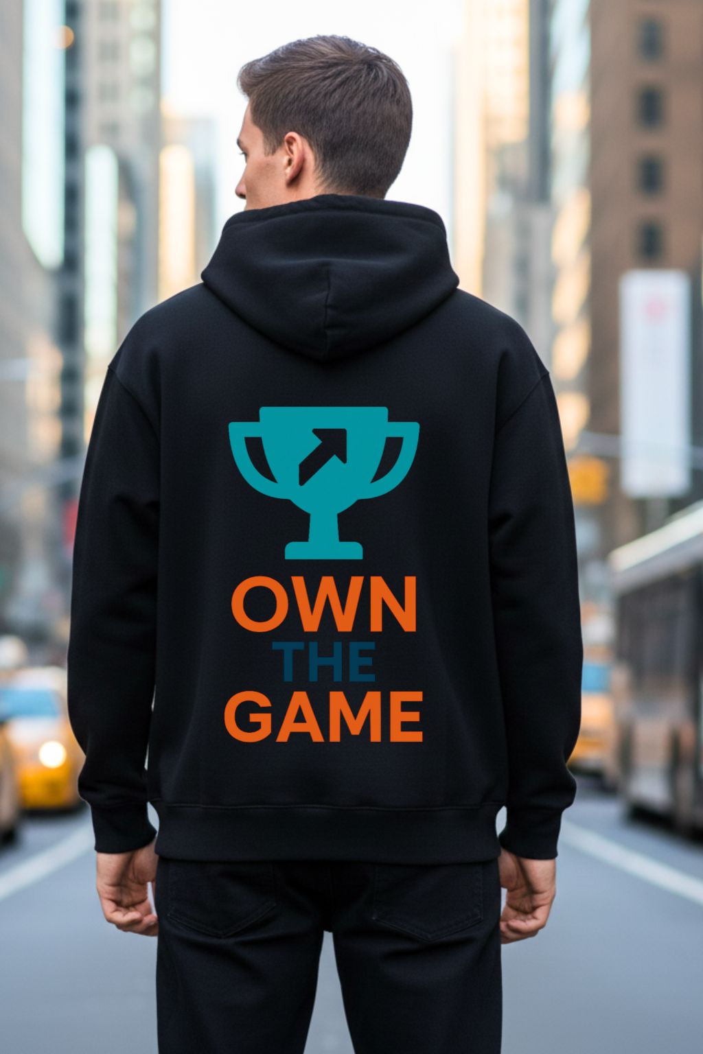 Back view of a man wearing a black oversized  hoodie with 'OWN THE GAME' text and logo on the back, standing in an urban setting.