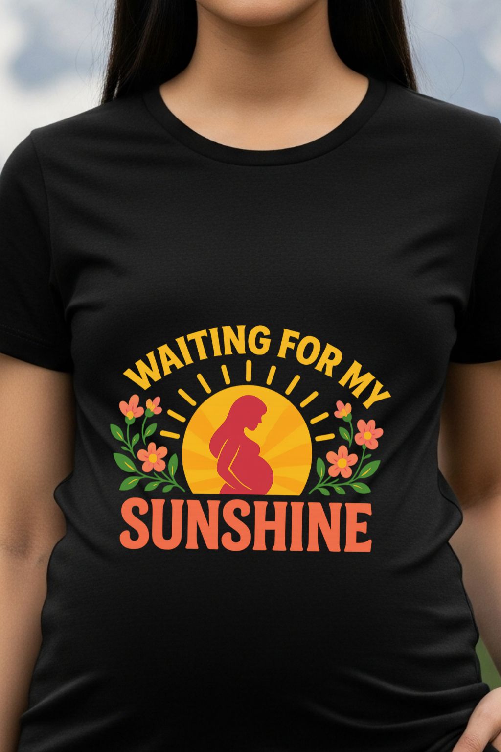 Woman wearing a Black maternity dress with a graphic design of a woman, sun, and flowers with text 'Waiting for my Sunshine' on a blurred natural background.